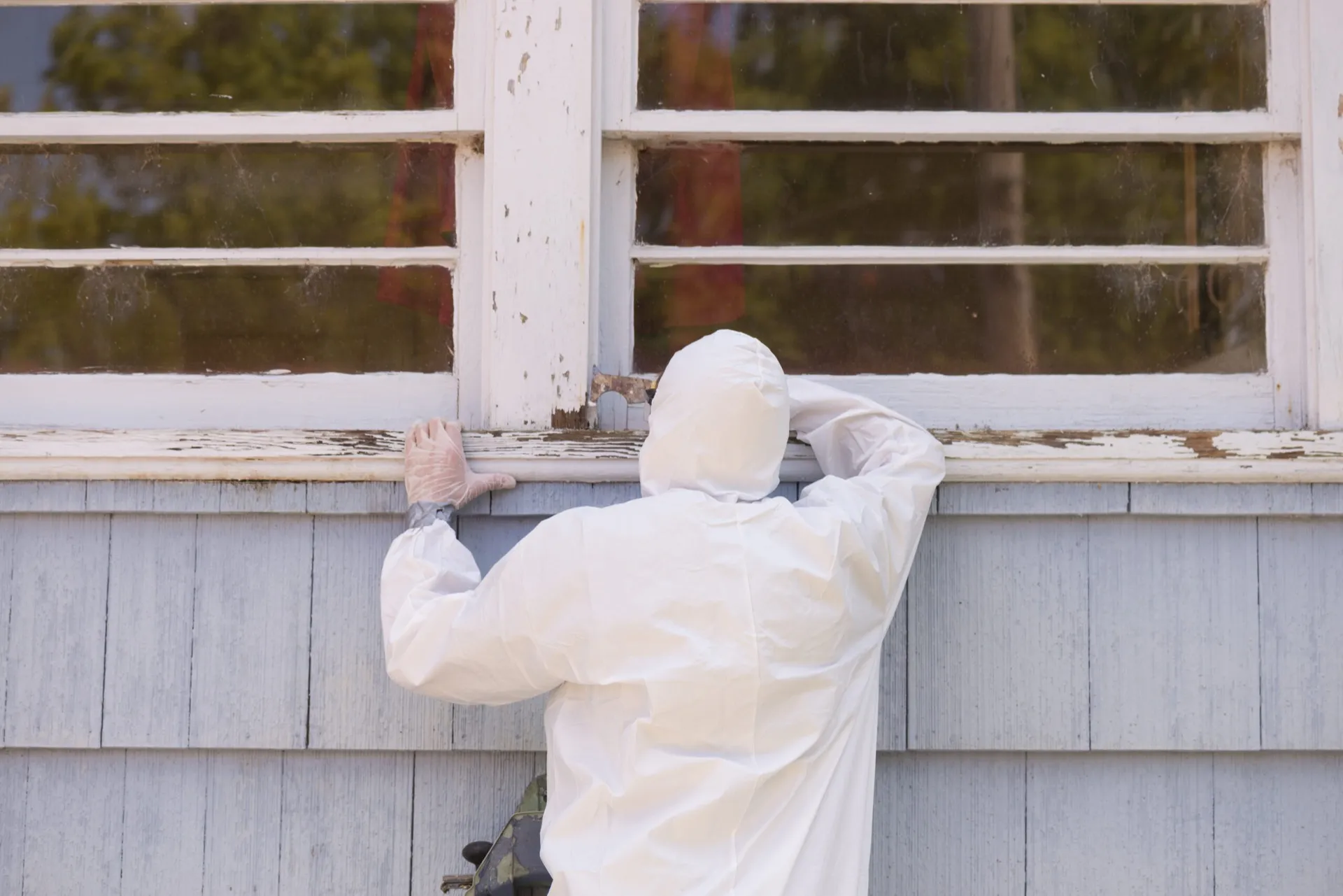 Certified professional in protective suit safely removing old lead paint from home siding