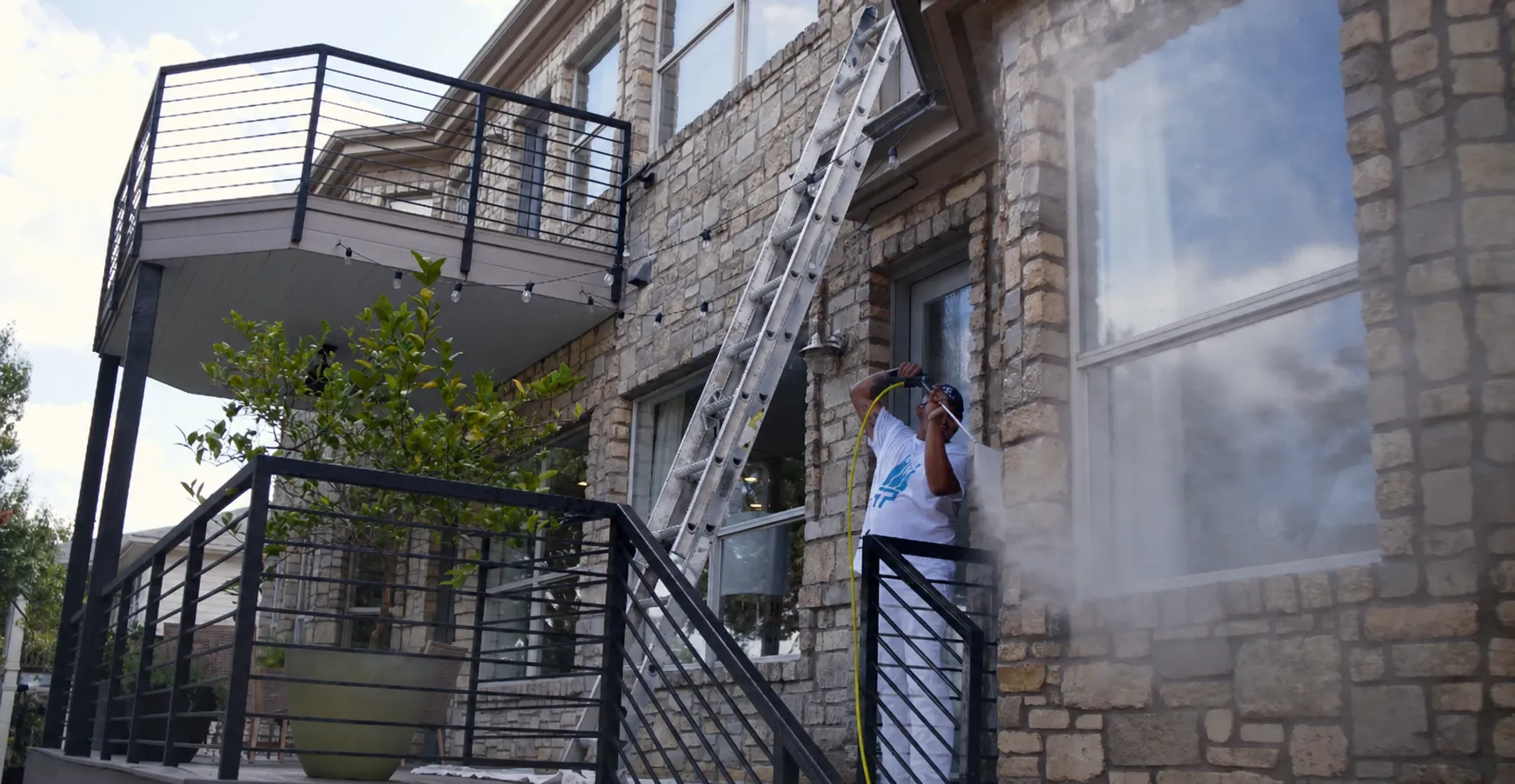 Painter on stairs pressure washing stone home exterior with ladder