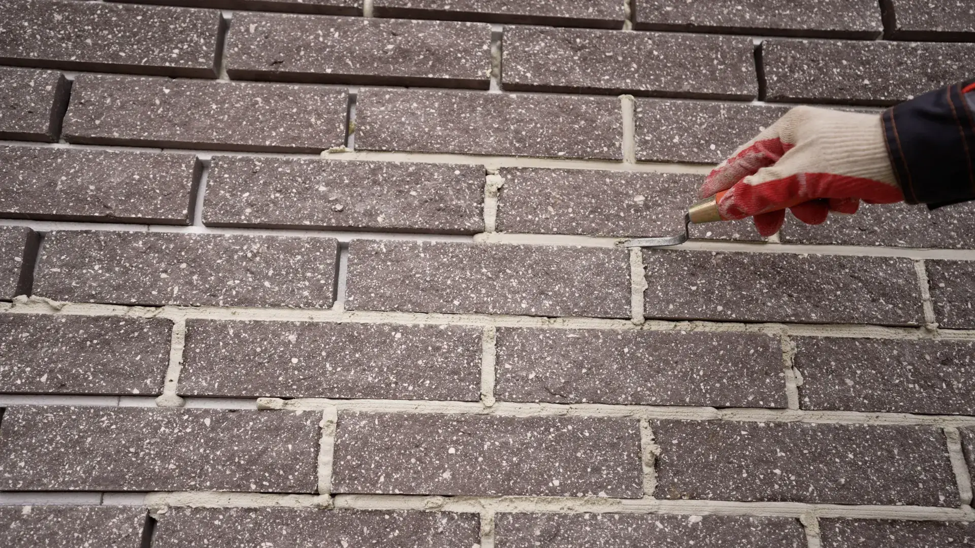 Gloved hand tuck-pointing fresh mortar between bricks on an exterior wall