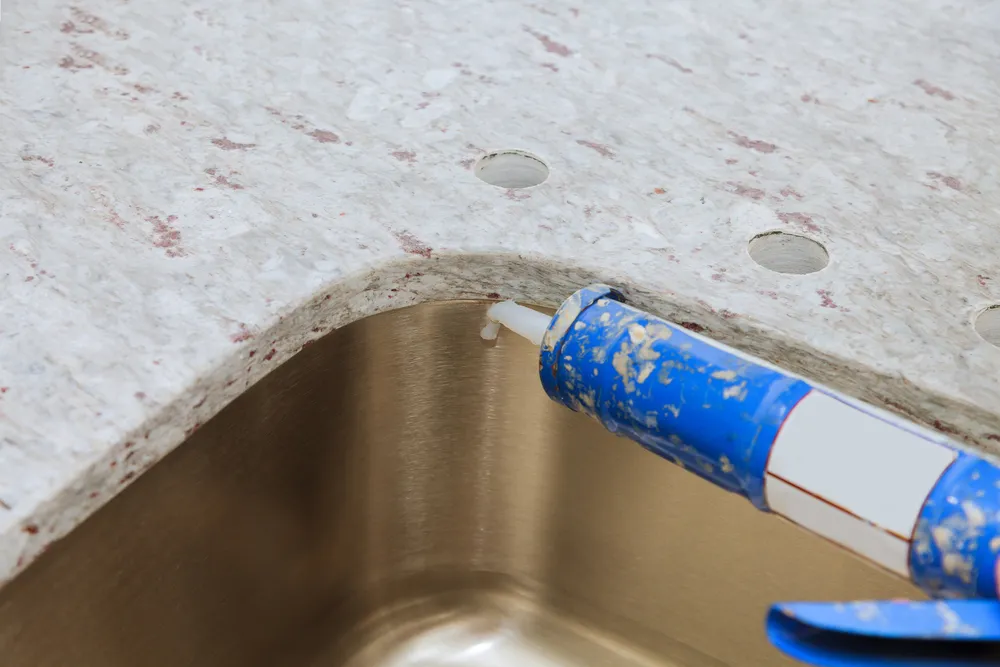 Fresh silicone caulk bead applied around a stainless kitchen sink and stone countertop
